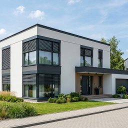 Firma handlowo-usługowa JAKUB RAJ - Nowoczesny dom jednorodzinny z szarymi żaluzjami zewnętrznymi na oknach, widok z zewnątrz, w tle inne domy i zieleń. Jasna elewacja, kostka brukowa.
