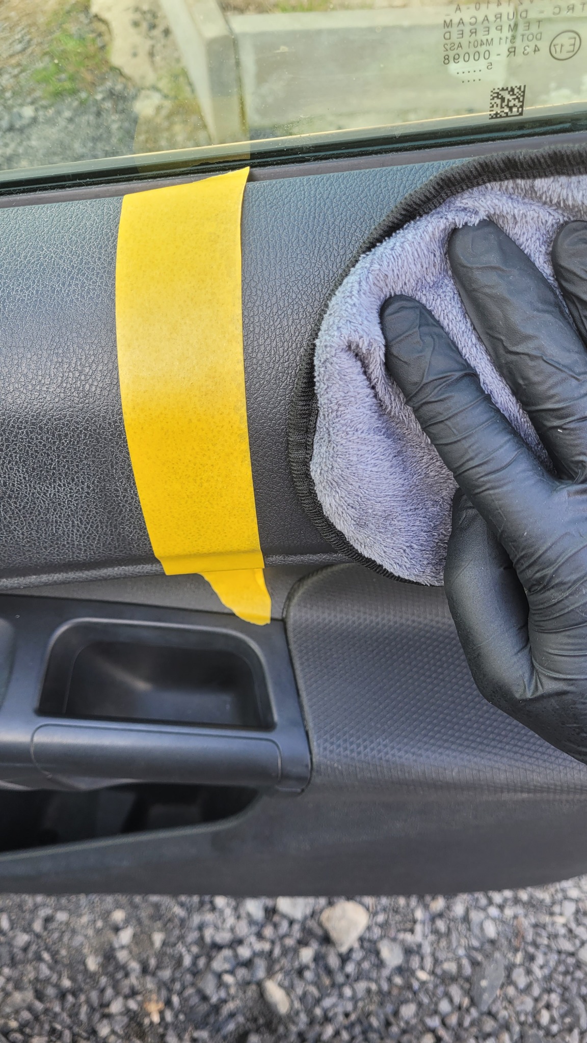 Czyszczenie wnętrza samochodu: ręka w czarnej rękawicy poleruje panel drzwiowy szarą ściereczką, obok żółta taśma maskująca. Detailing samochodowy w trakcie.