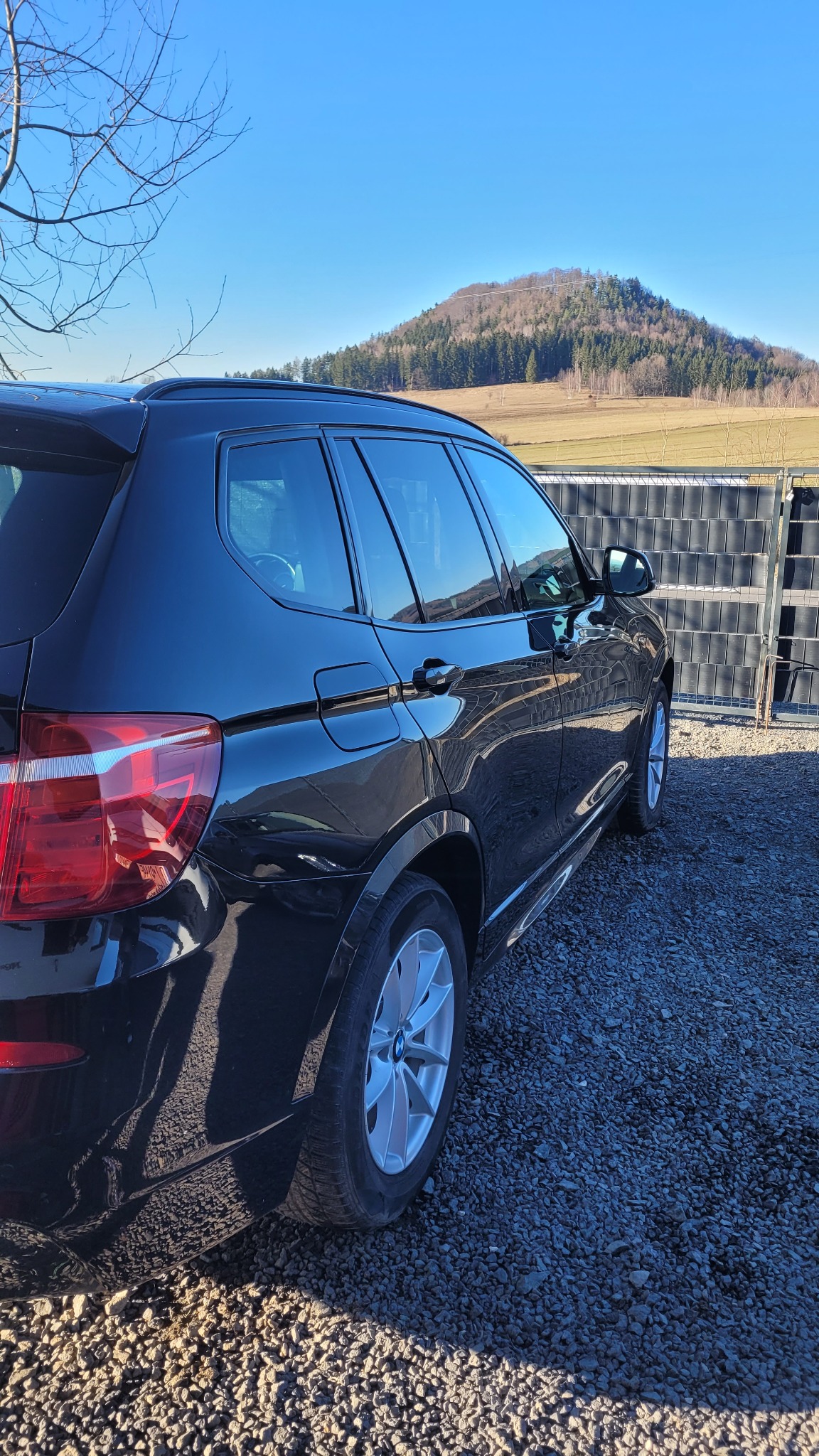 Błyszczący, czarny SUV BMW X3 zaparkowany na żwirowym podłożu, z widokiem na malownicze wzgórze w tle pod błękitnym niebem. Widoczny fragment ogrodzenia.