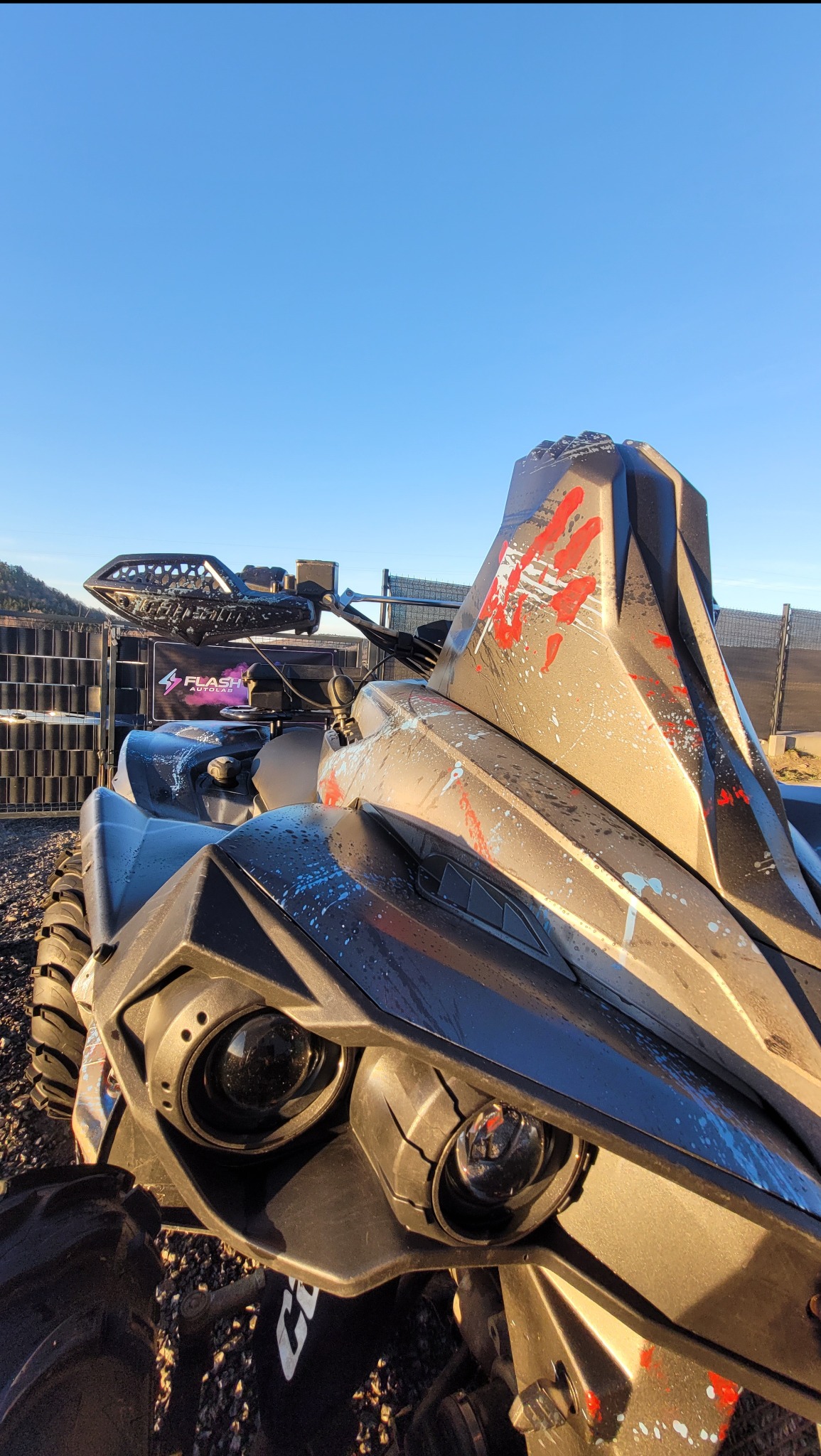 Umyty, czarny quad z czerwonymi i białymi plamami farby, stojący na żwirowym podłożu w słoneczny dzień. Widoczne opony i reflektory.