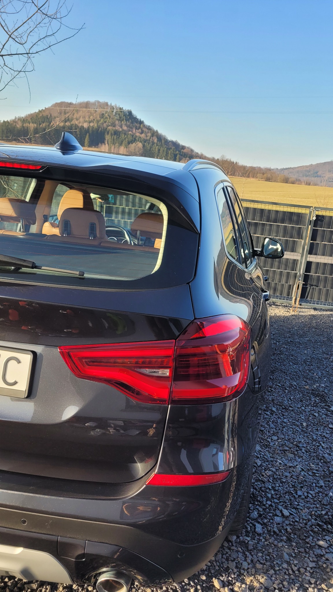 Ciemny SUV, widok z tyłu, zarys gór w tle. Widoczne detale: światła, tablica rejestracyjna, wycieraczka. Auto stoi na żwirowym podłożu, obok metalowe ogrodzenie.