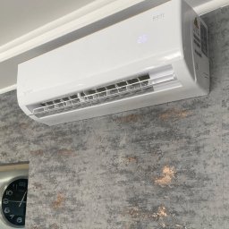 Wentica Tomasz Manowski - Biały klimatyzator Roni zamontowany na szarej ścianie o teksturze betonu. Widoczny fragment zegara w lewym dolnym rogu. Wyświetlacz klimatyzatora pokazuje temperaturę 26 stopni.