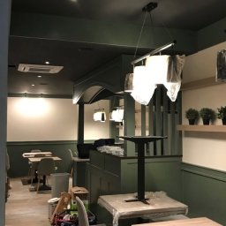 Wentica Tomasz Manowski - Wnętrze lokalu komercyjnego w trakcie montażu klimatyzacji. Widoczny klimatyzator na suficie, stoliki z krzesłami i elementy dekoracyjne w odcieniach zieleni i bieli.