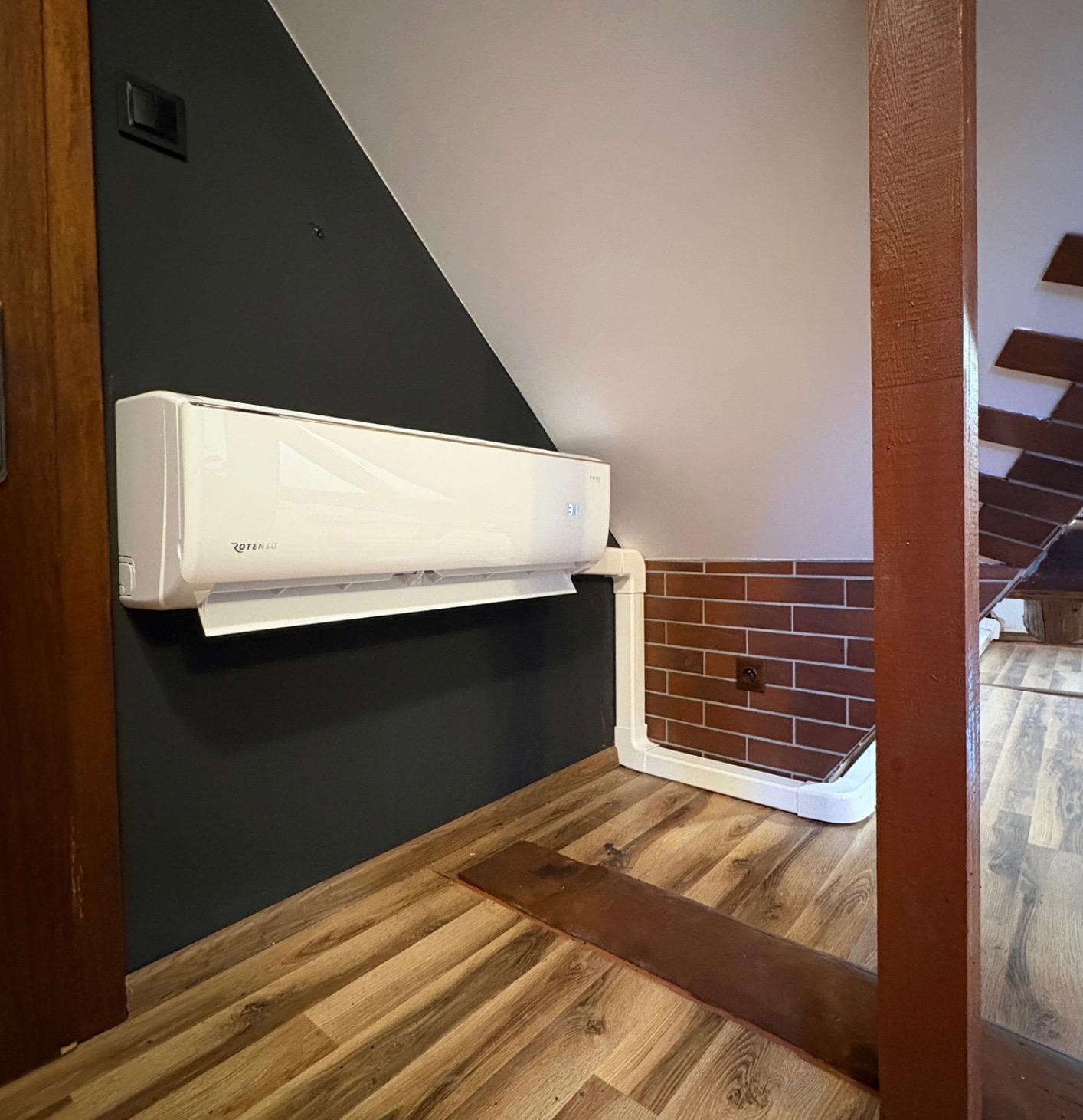 Biały klimatyzator Rotenso zamontowany na skośnej, ciemnej ścianie poddasza, z widocznymi białymi rurami instalacyjnymi i drewnianą podłogą.