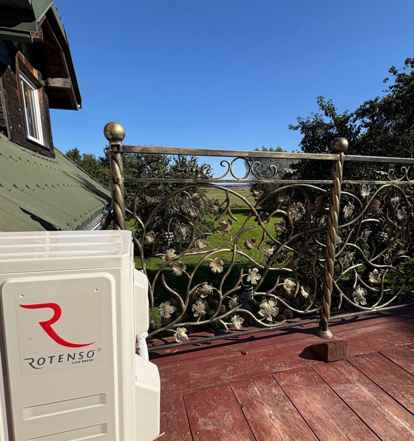Biała jednostka zewnętrzna klimatyzacji Rotenso na drewnianym tarasie z ozdobną, metalową balustradą na tle zieleni i błękitnego nieba.
