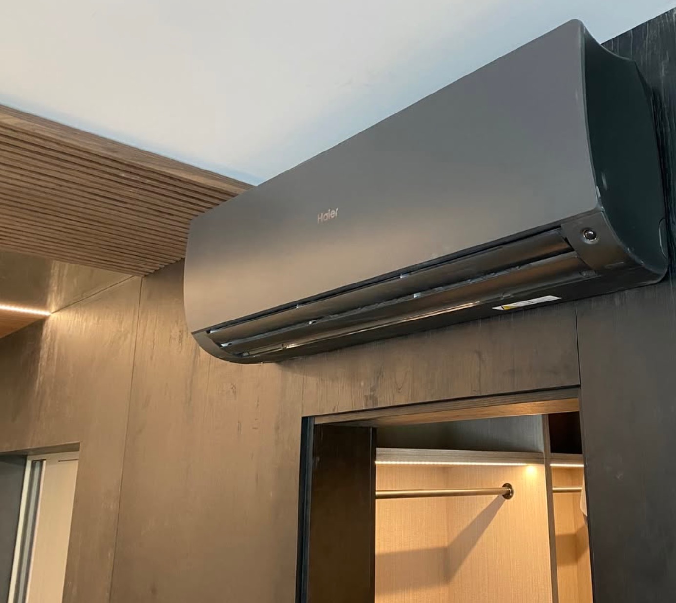 Zamontowany, grafitowy klimatyzator Haier w nowoczesnym wnętrzu z drewnianymi akcentami i oświetleniem LED. Widok z dołu, podkreślający minimalistyczny design.