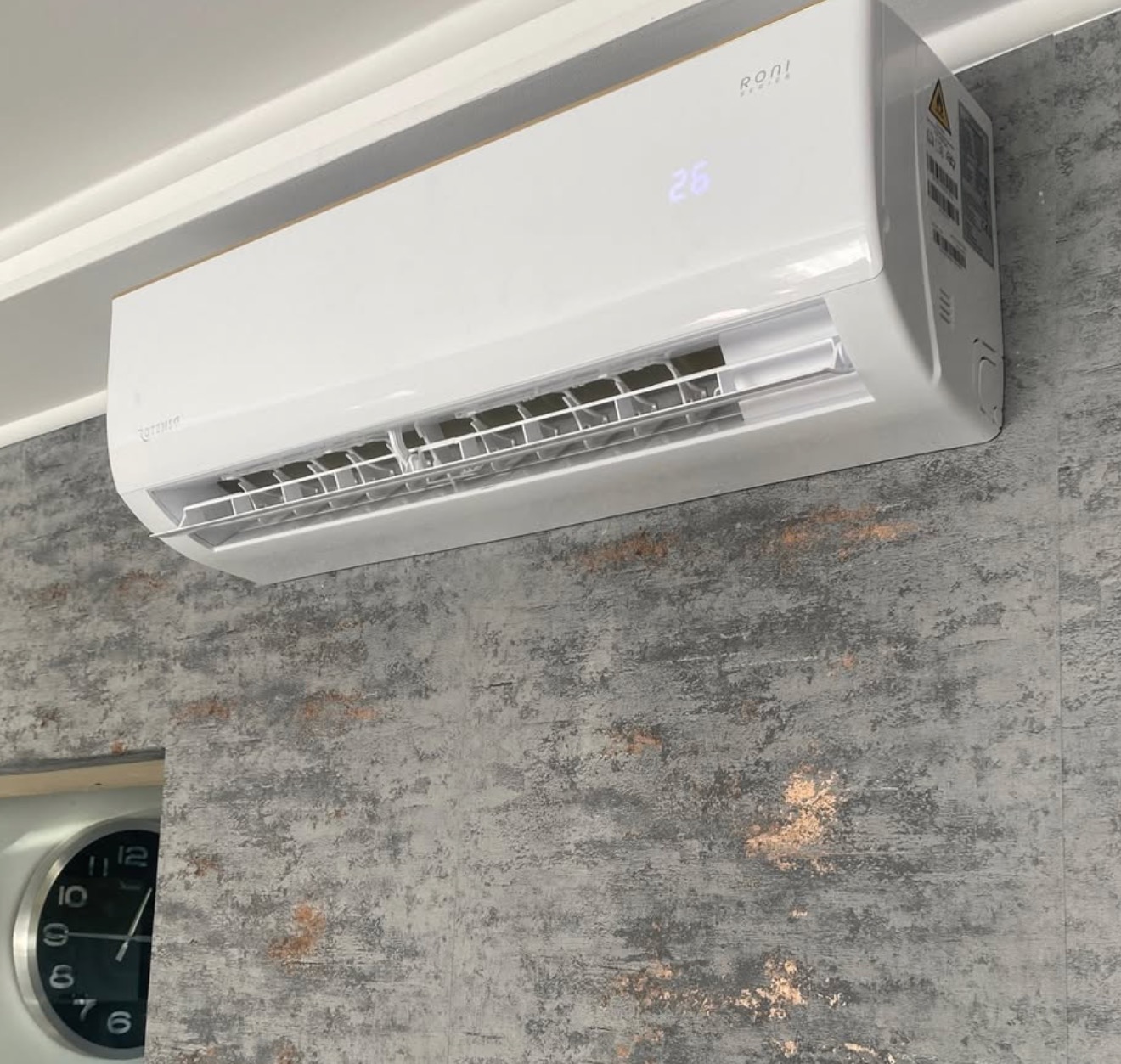 Biały klimatyzator Roni zamontowany na szarej ścianie o teksturze betonu. Widoczny fragment zegara w lewym dolnym rogu. Wyświetlacz klimatyzatora pokazuje temperaturę 26 stopni.