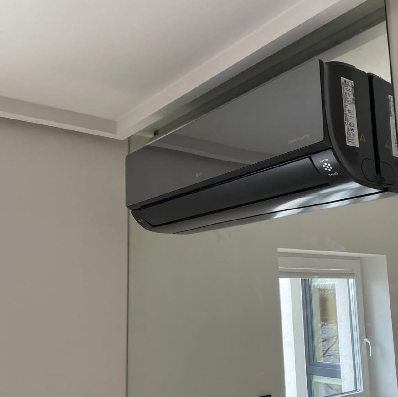 Czarny klimatyzator LG Dual Inverter zamontowany pod sufitem, odbijający się w lustrze. Widoczne okno w tle. Minimalistyczny design wnętrza.