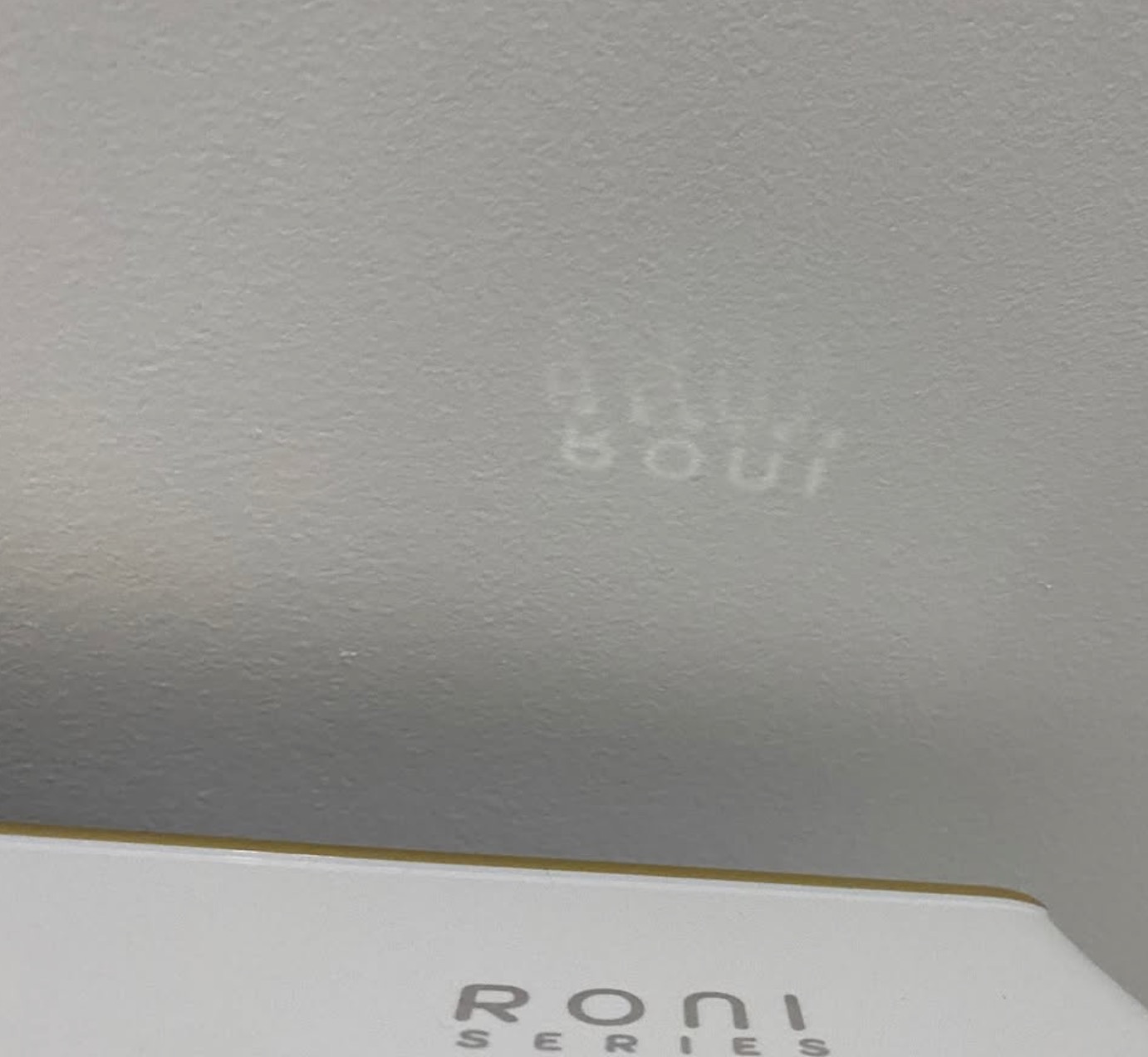 Biała ściana z delikatnym cieniem napisu 'RONI SERIES' rzucanym przez urządzenie, prawdopodobnie klimatyzacyjne lub wentylacyjne.