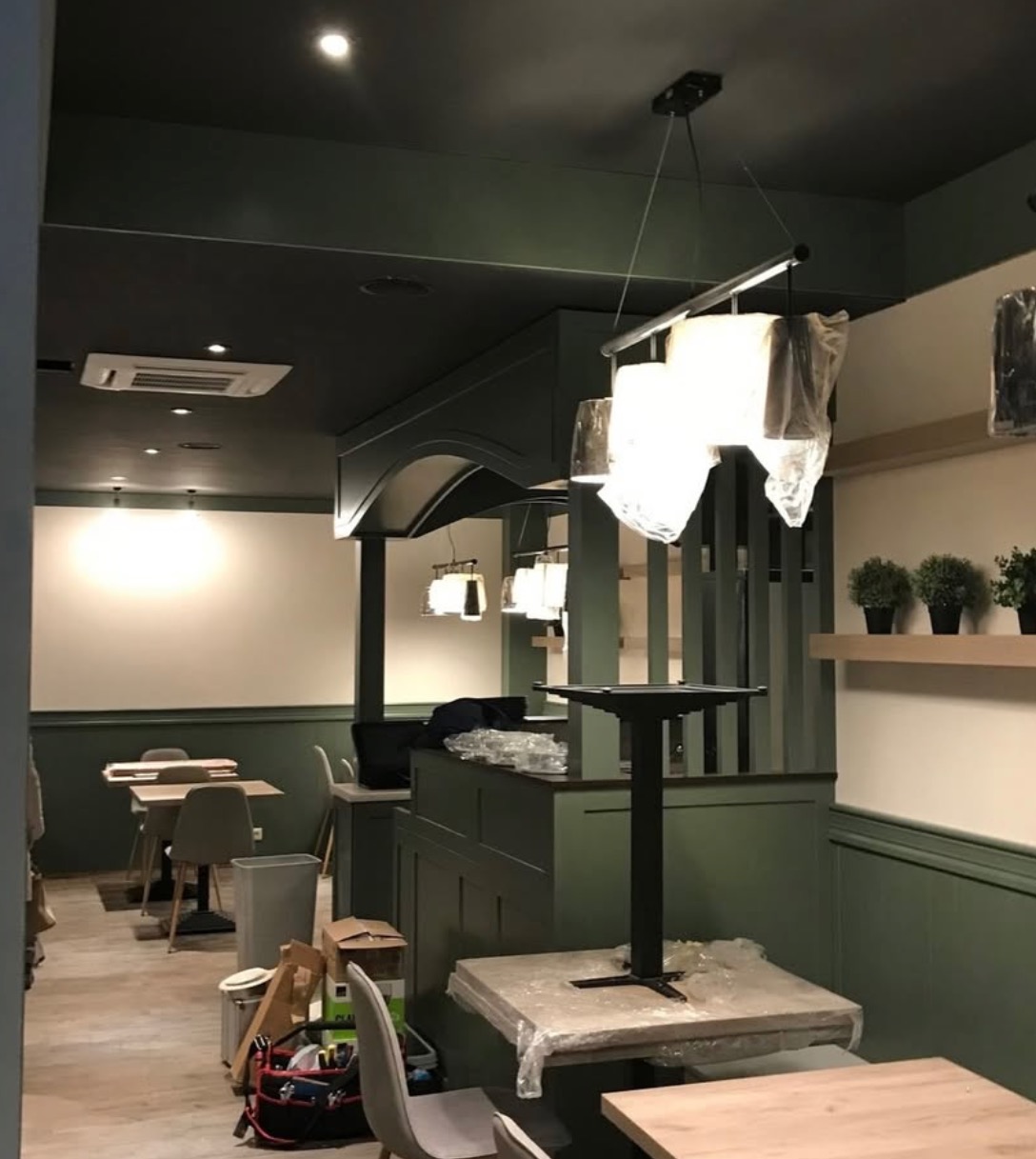 Wnętrze lokalu komercyjnego w trakcie montażu klimatyzacji. Widoczny klimatyzator na suficie, stoliki z krzesłami i elementy dekoracyjne w odcieniach zieleni i bieli.