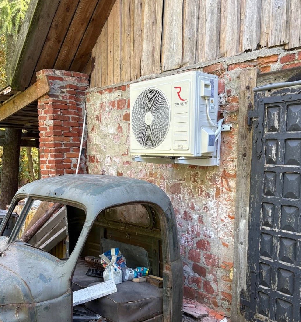 Biały klimatyzator Rotenso zamontowany na ceglanej ścianie budynku gospodarczego, obok stoi stary, zardzewiały samochód dostawczy bez karoserii.