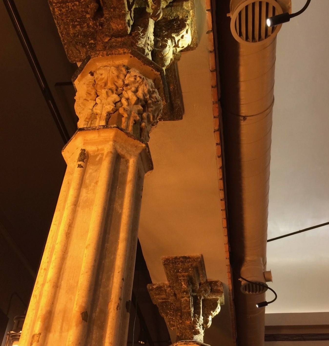 Antyczna kolumna z dekoracyjną głowicą sąsiaduje z nowoczesną rurą wentylacyjną pod sufitem, oświetloną punktowym światłem. Kontrast stylów i epok.