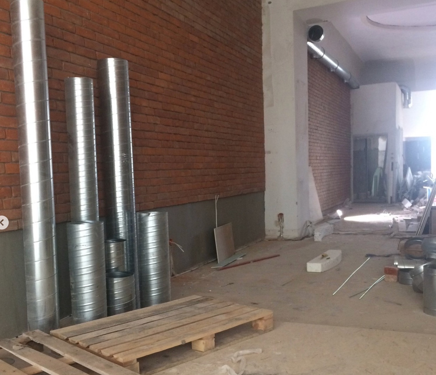 Stalowe rury wentylacyjne ułożone przy ceglanej ścianie w surowym wnętrzu budynku w trakcie montażu instalacji. Widoczne palety i elementy instalacji.