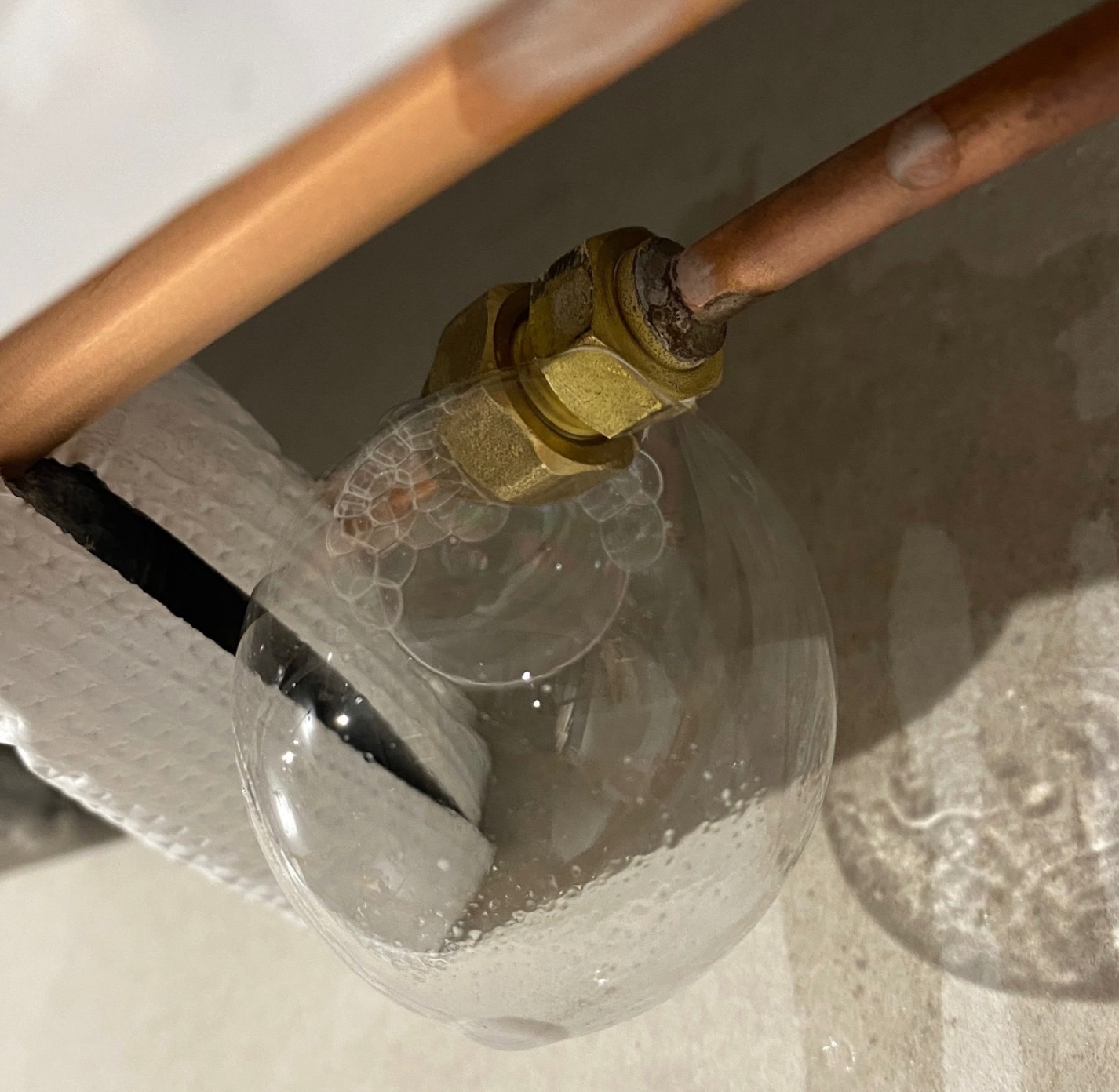 Wyciek z miedzianej rury, testowany roztworem mydła. Widoczne pęcherzyki powietrza wskazują nieszczelność na złączce. Biała szmatka w tle.