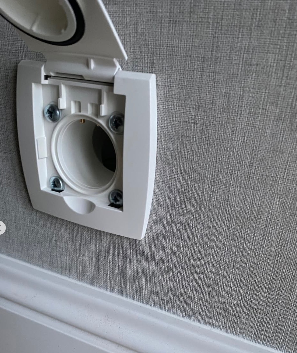 Białe gniazdo ssące centralnego odkurzacza z otwartą klapką, zamontowane na szarej ścianie z widoczną listwą przypodłogową. Widoczne śruby mocujące.