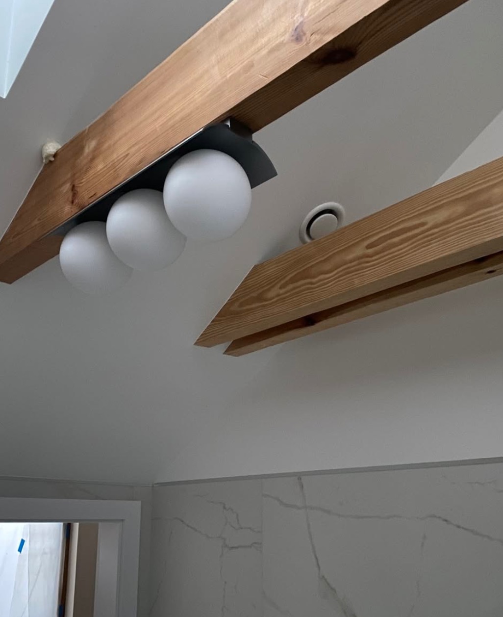 Nowoczesna łazienka z drewnianymi belkami i okrągłymi lampami sufitowymi. Widoczny fragment wentylacji sufitowej i marmurowe płytki na ścianach.