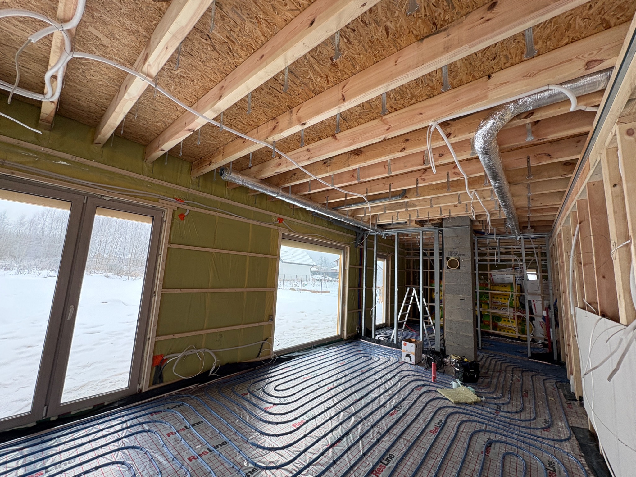 Wnętrze w trakcie montażu instalacji: rury ogrzewania podłogowego, izolacja ścian, elementy wentylacji pod sufitem. Widok na zasypany śniegiem krajobraz za oknem.
