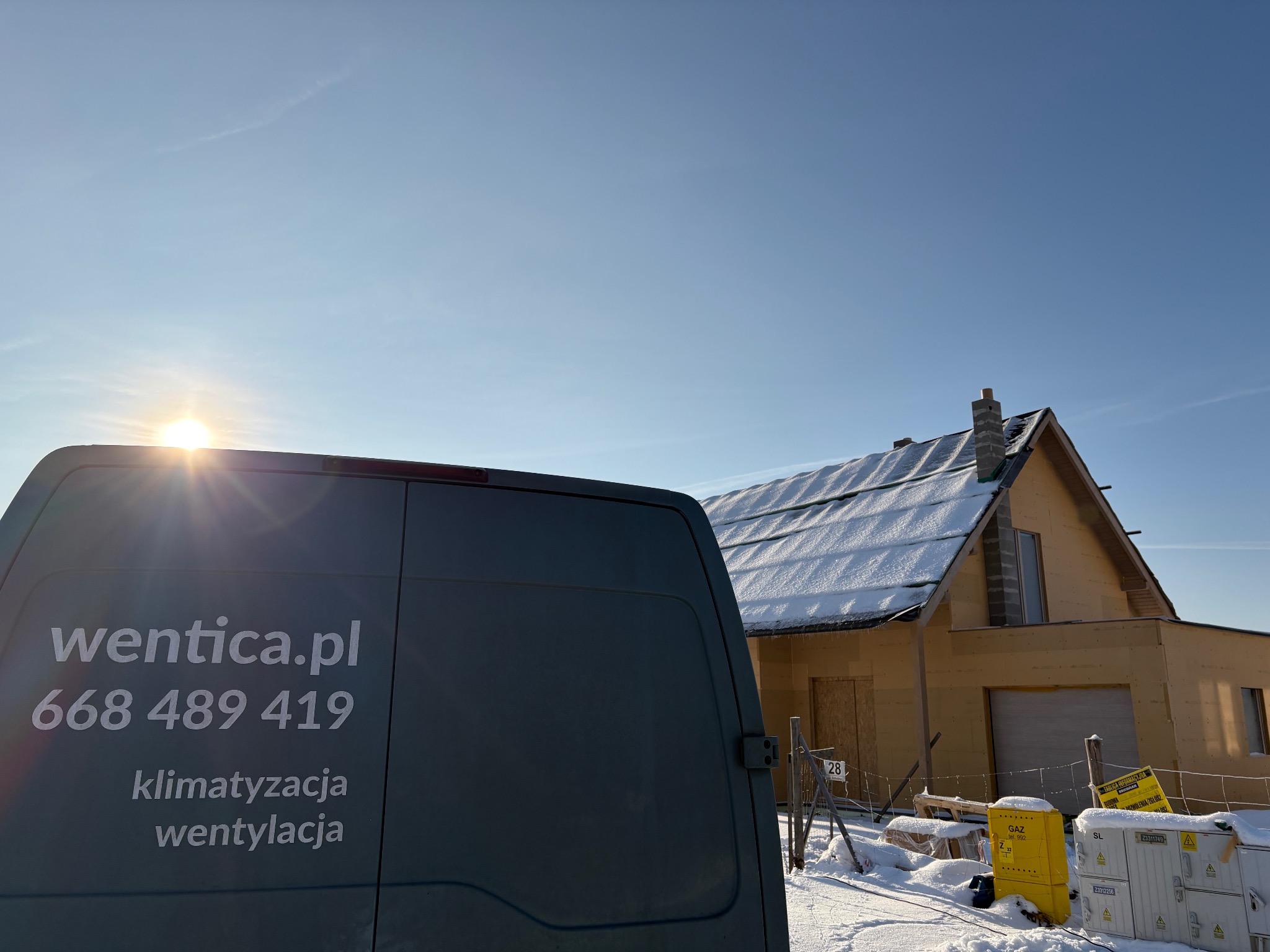 Szary samochód dostawczy z logo wentica.pl i numerem telefonu na tle ośnieżonego domu w budowie. Widoczne skrzynki gazowe i elementy instalacji.