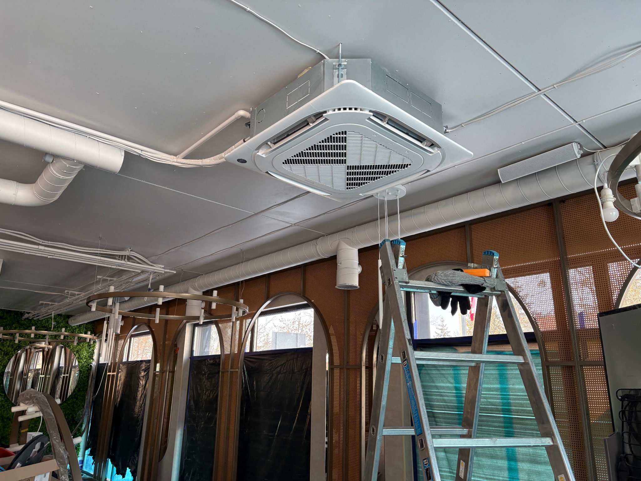 Sufitowa klimatyzacja kasetonowa w jasnym wnętrzu, widoczna drabina i instalacje. Ujęcie z dołu, podkreślające nowoczesny design urządzenia.