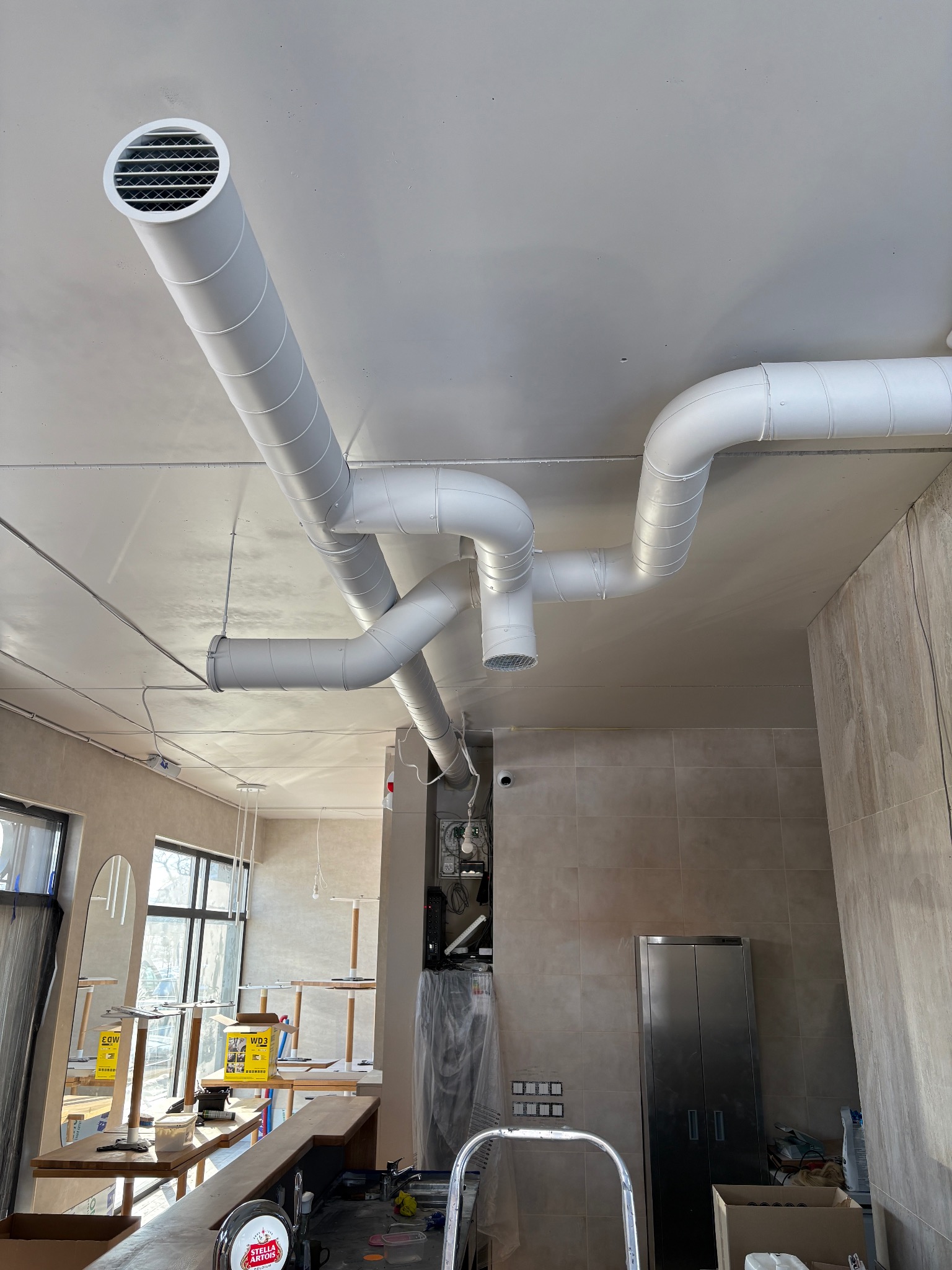Białe rury wentylacyjne pod sufitem w lokalu komercyjnym, widoczne kratki wylotowe, surowy stan wykończenia wnętrza, płytki na ścianach.