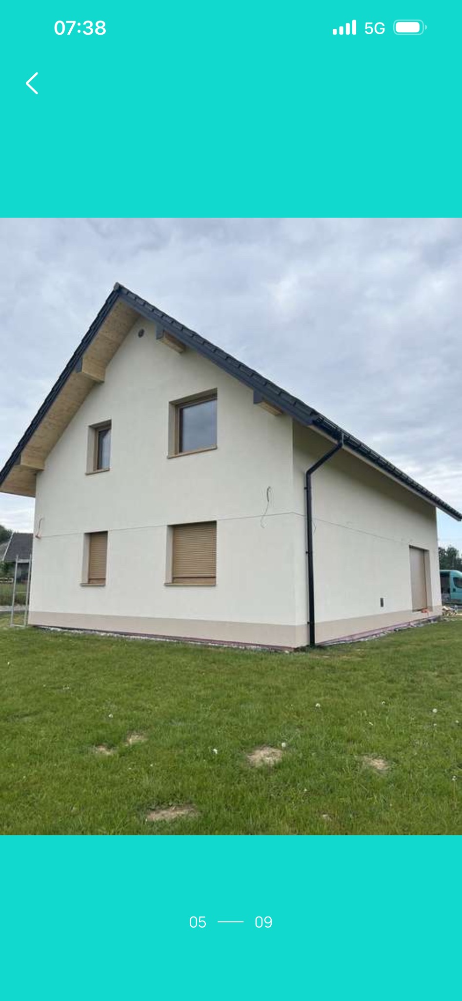 Dom jednorodzinny z jasną elewacją i ciemnym dachem, widoczny z poziomu trawnika. Okna z roletami, czarne rynny. Świeżo wykończona elewacja.