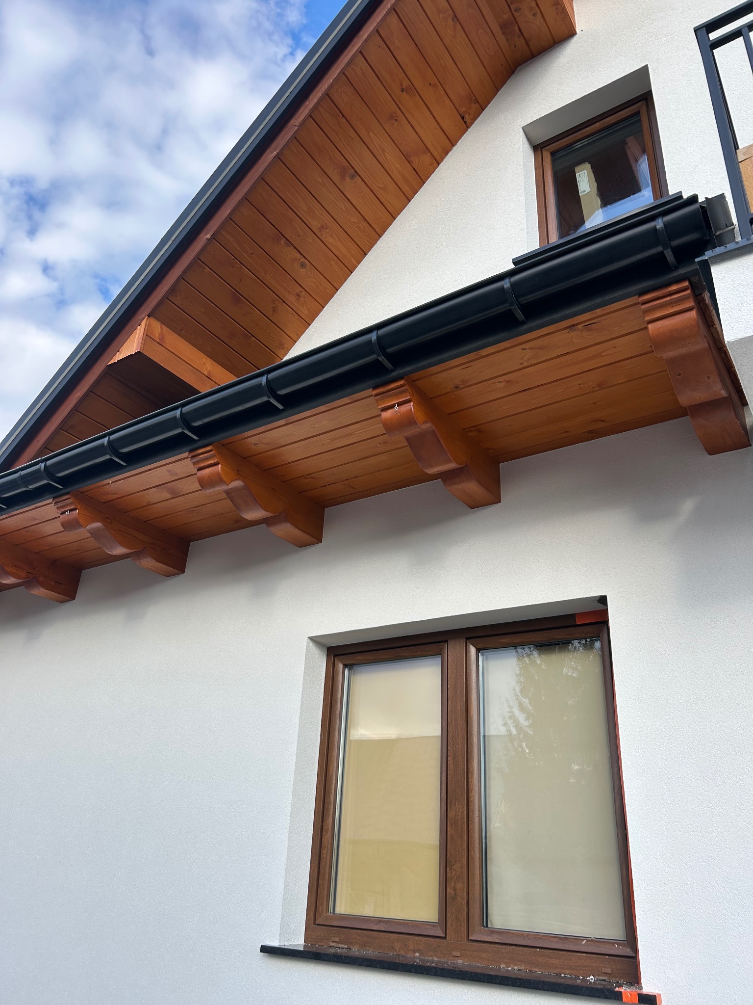 Biała elewacja domu z drewnianym okapem i czarnymi rynnami. Widoczne okna z brązowymi ramami. Ujęcie z dołu, podkreślające detale wykończenia.