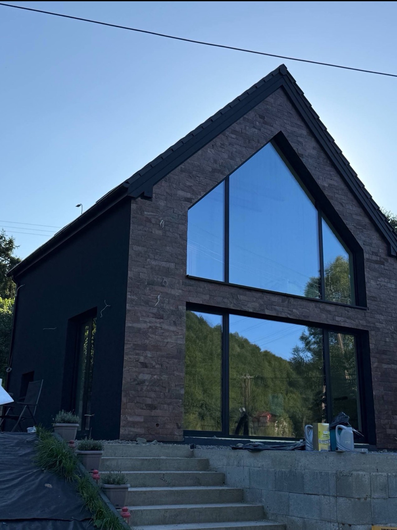 Nowoczesny dom z elewacją z kamienia i dużymi oknami, odbijającymi krajobraz górski. Część elewacji pomalowana na czarno. Widoczne schody wejściowe i elementy wykończeniowe.