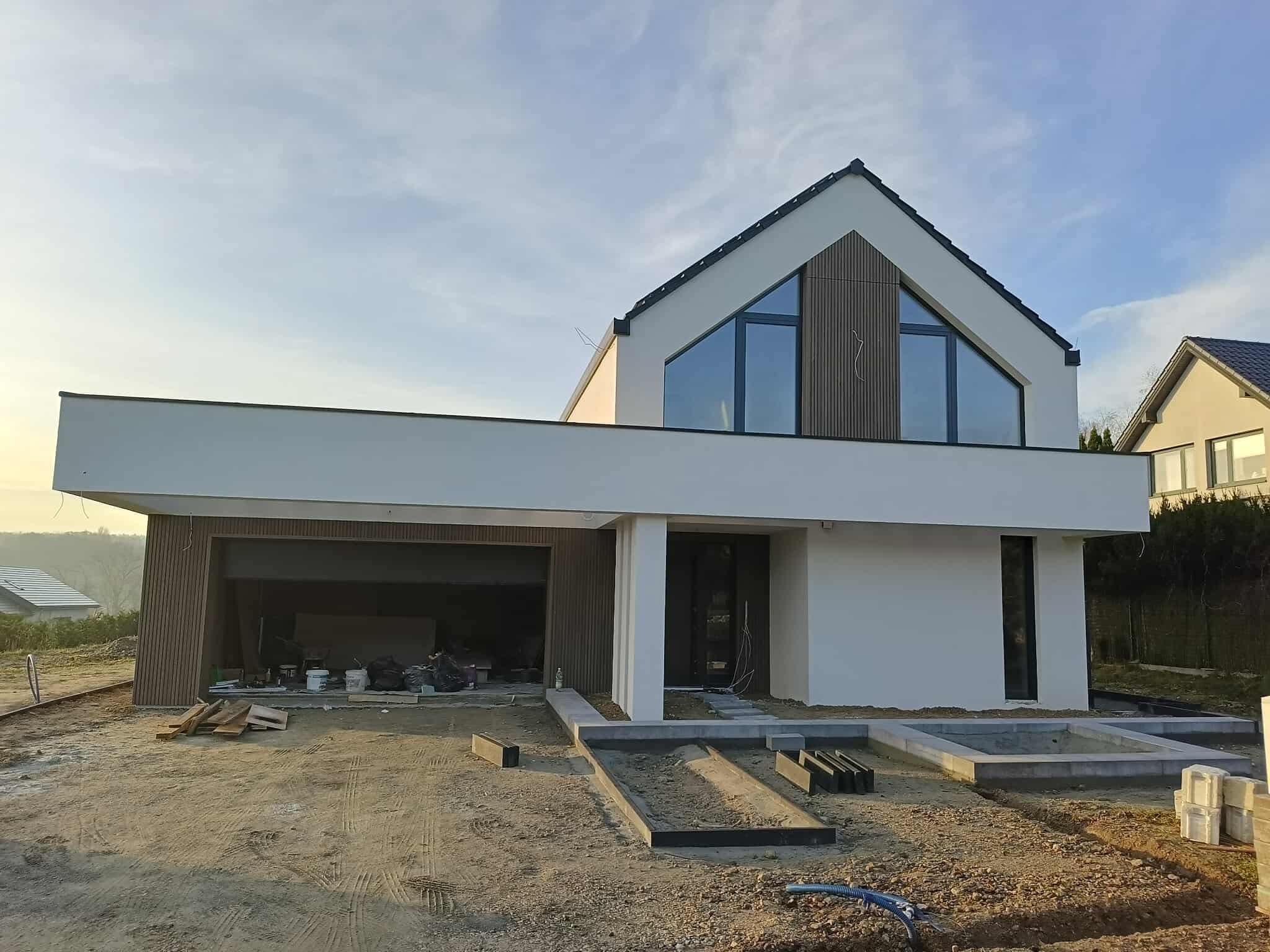 Nowoczesny dom jednorodzinny z jasną elewacją i elementami drewnianymi, w trakcie wykańczania. Widoczne przygotowania do zagospodarowania terenu.