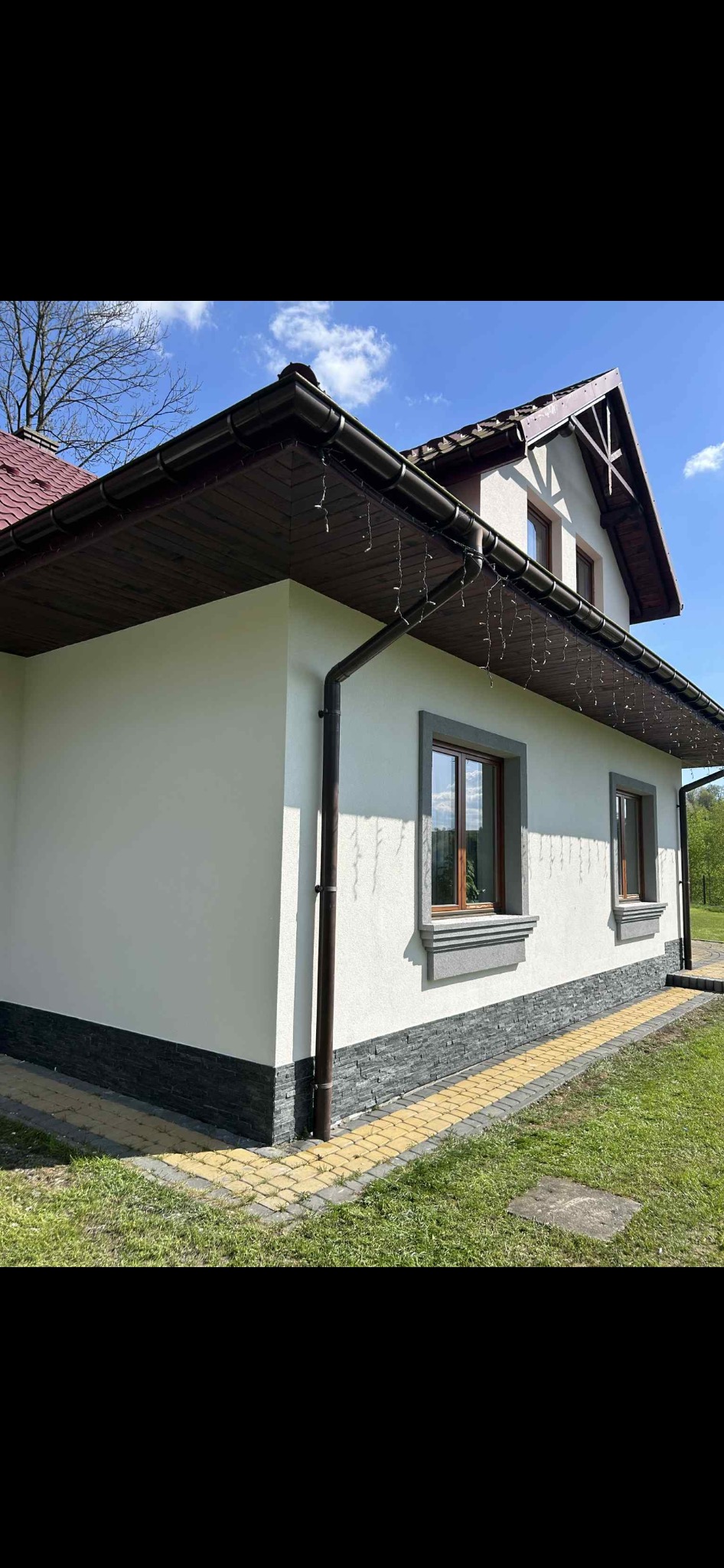 Wykończona elewacja domu jednorodzinnego z białym tynkiem, ciemną podmurówką z kamienia i brązowymi rynnami. Widoczne okna z szarymi obramowaniami i fragment dachu.