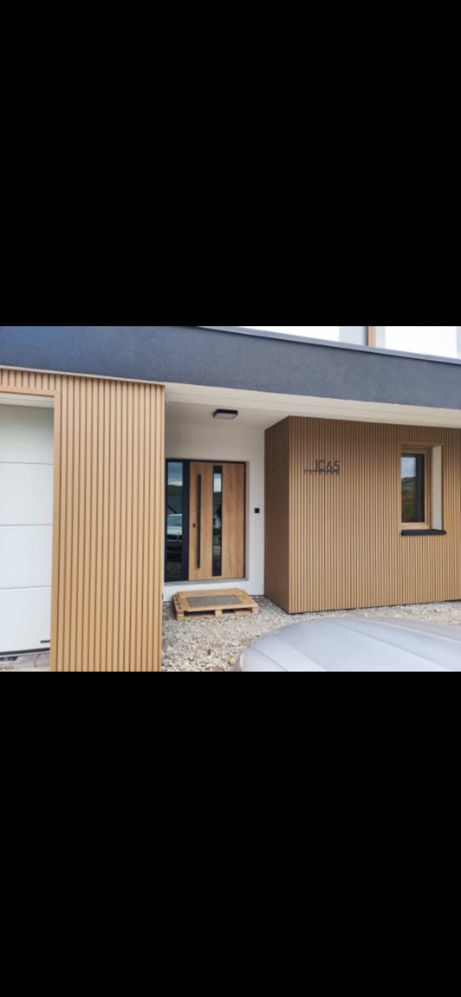 Nowoczesny dom z elewacją z drewnianych paneli pionowych, numer domu '1065' nad oknem. Wejście z drewnianymi drzwiami i szarym podłożem.