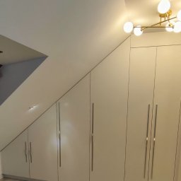 Karol Dyduch - Biała szafa wnękowa z długimi, srebrnymi uchwytami, dopasowana do skosu poddasza. Jasna podłoga i nowoczesna lampa sufitowa.