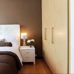 Karol Dyduch - Nowoczesna sypialnia z beżową szafą przesuwną, białym łóżkiem z pikowanym zagłówkiem i stolikiem nocnym z lampką. W tle widoczny fragment innego pomieszczenia.