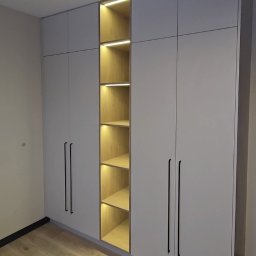 Karol Dyduch - Jasnoszara szafa wnękowa z oświetloną wnęką na półki, czarne, długie uchwyty, drewniana podłoga. Minimalistyczny design.