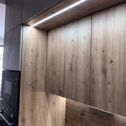 Karol Dyduch - Drewniane fronty szafek kuchennych z oświetleniem LED, widoczny fragment zabudowy z piekarnikiem. Ujęcie z bliska, ukazujące strukturę drewna i detale wykończenia.