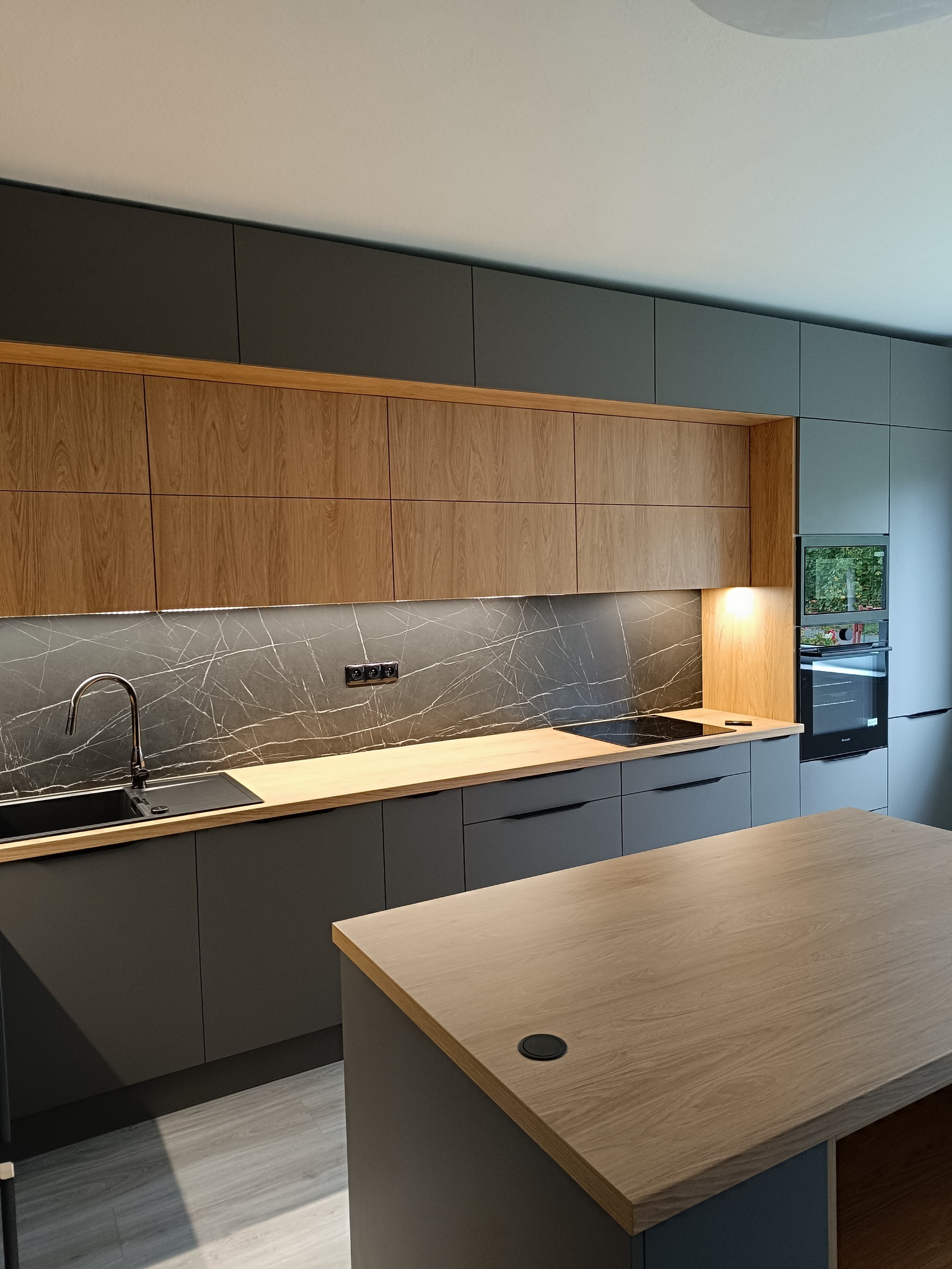Nowoczesna kuchnia z szarymi szafkami dolnymi i drewnianymi górnymi, marmurowym blatem i wyspą. Minimalistyczny design z oświetleniem LED pod szafkami.