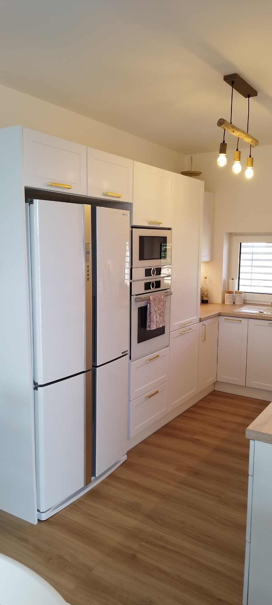 Biała kuchnia na wymiar z lodówką side-by-side, piekarnikiem i mikrofalówką w zabudowie. Złote uchwyty i drewniana lampa dodają ciepła. Jasne drewno na podłodze.