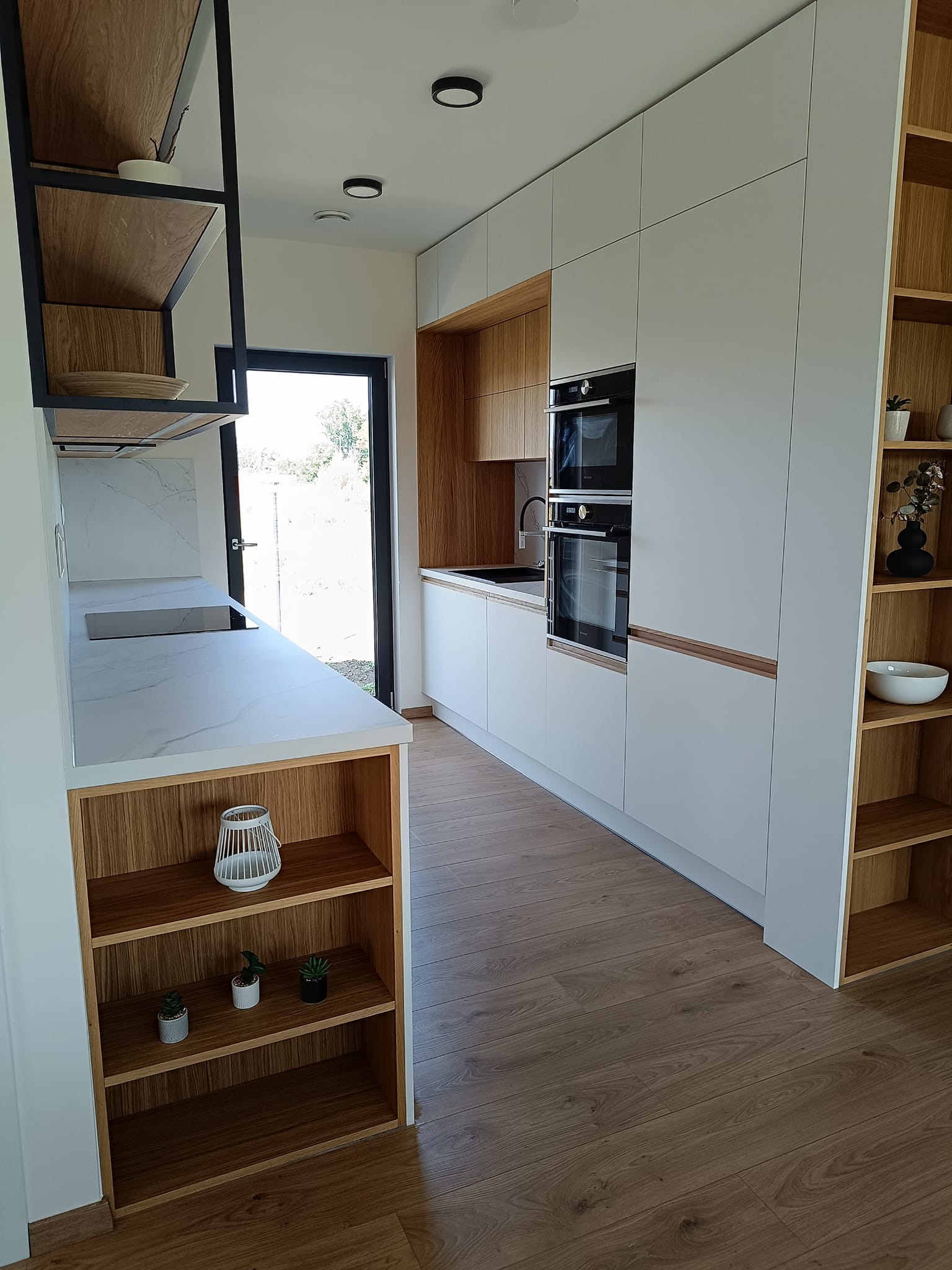 Nowoczesna kuchnia z białymi frontami i drewnianymi akcentami. Widoczne półki z dekoracjami, blaty z marmurowym wzorem i zabudowane sprzęty AGD. Minimalistyczny design.