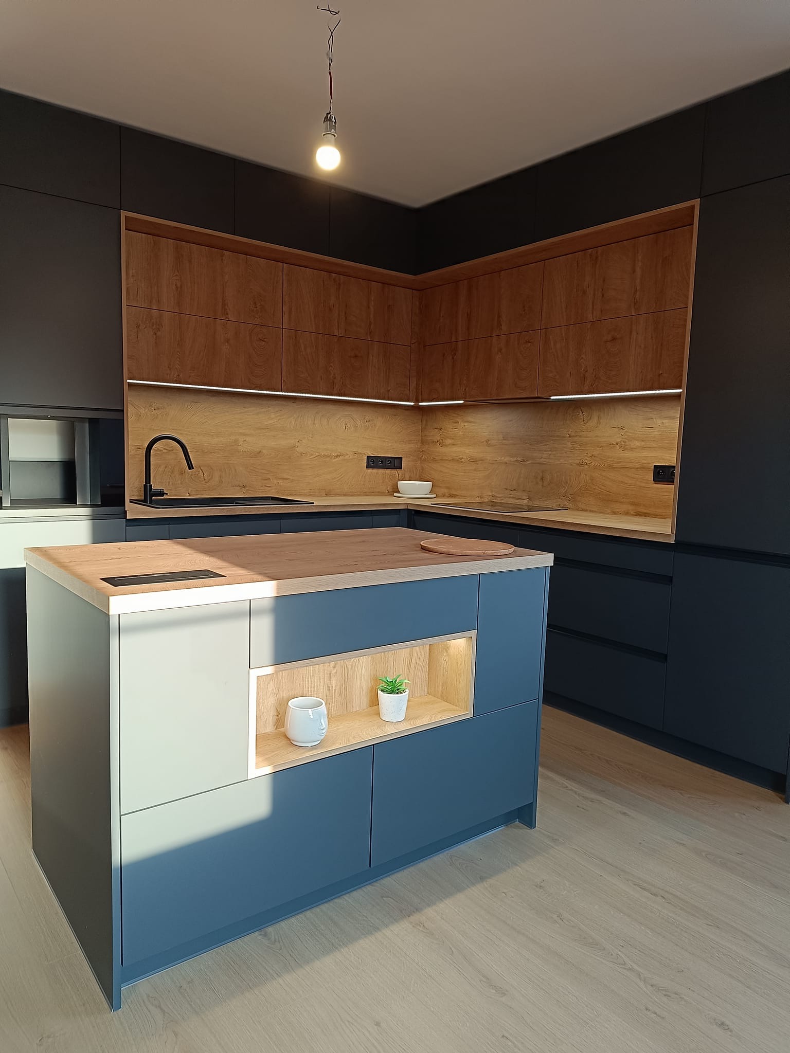 Nowoczesna kuchnia z wyspą: grafitowe szafki, drewniane akcenty, podświetlenie LED. Na wyspie dekoracje: roślina i kubek. Minimalistyczny design, ciepłe światło.
