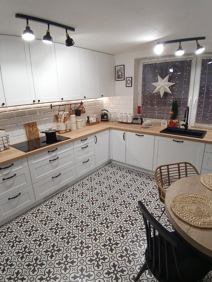 Biała kuchnia w stylu skandynawskim z drewnianym blatem, czarnymi uchwytami i wzorzystą podłogą. Dekoracje świąteczne przy oknie dodają wnętrzu przytulności.