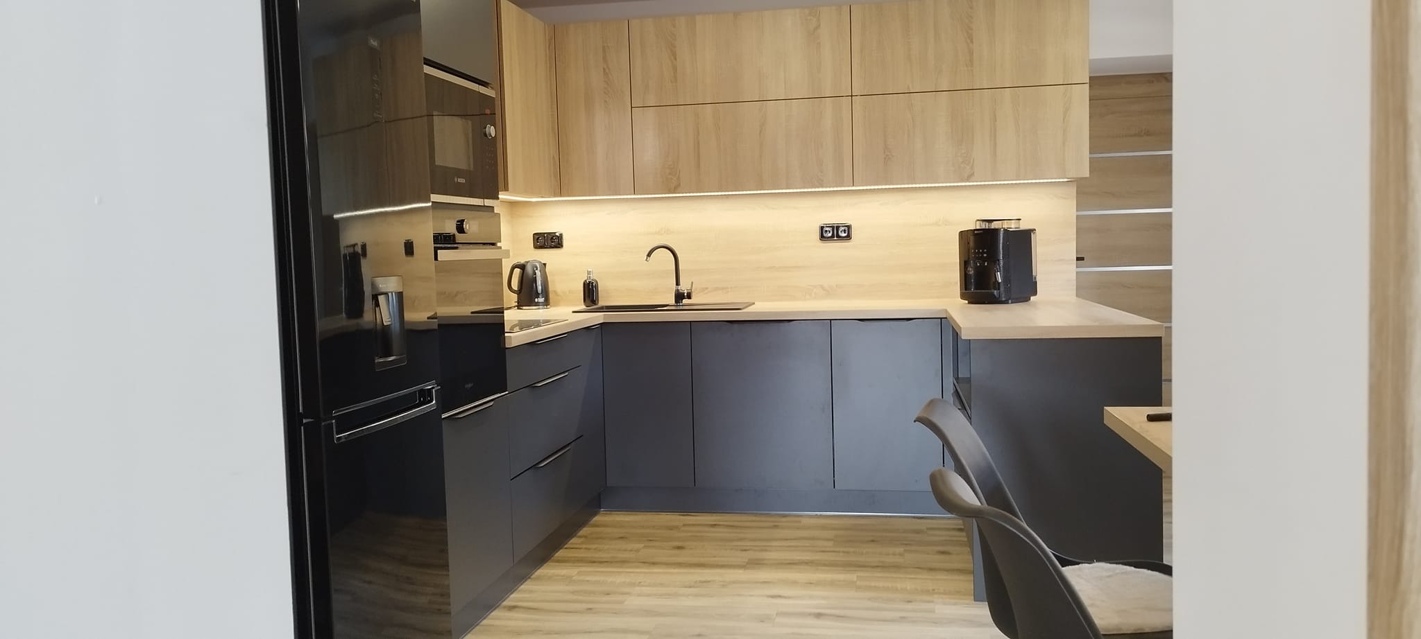 Nowoczesna kuchnia w Choczni z szarymi szafkami dolnymi i drewnopodobnymi górnymi, oświetlona listwą LED pod szafkami. Widoczny czarny sprzęt AGD.