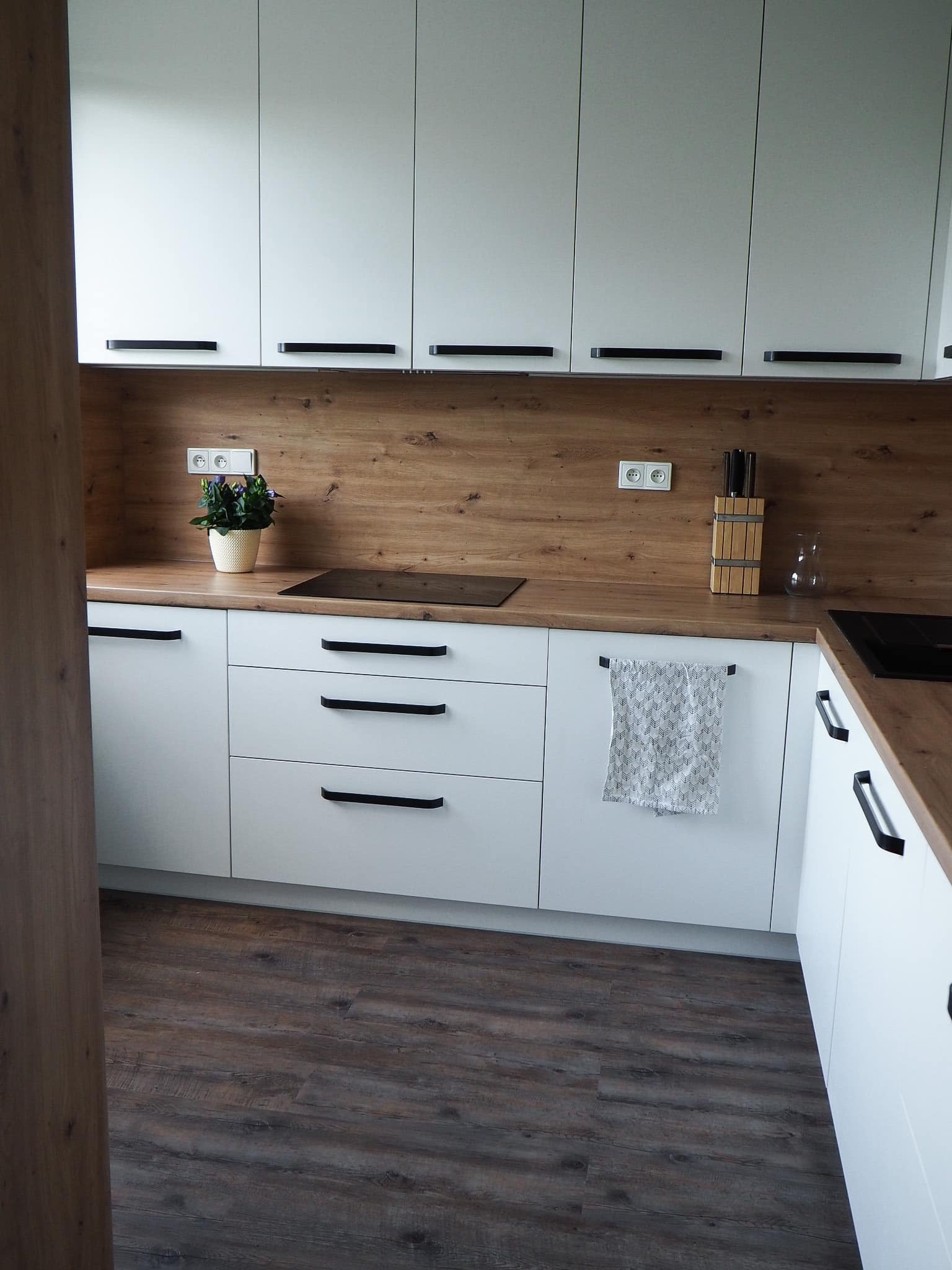Nowoczesna kuchnia z białymi frontami i drewnianym blatem. Minimalistyczne czarne uchwyty, płyta indukcyjna i akcent roślinny w postaci doniczki z kwiatami.
