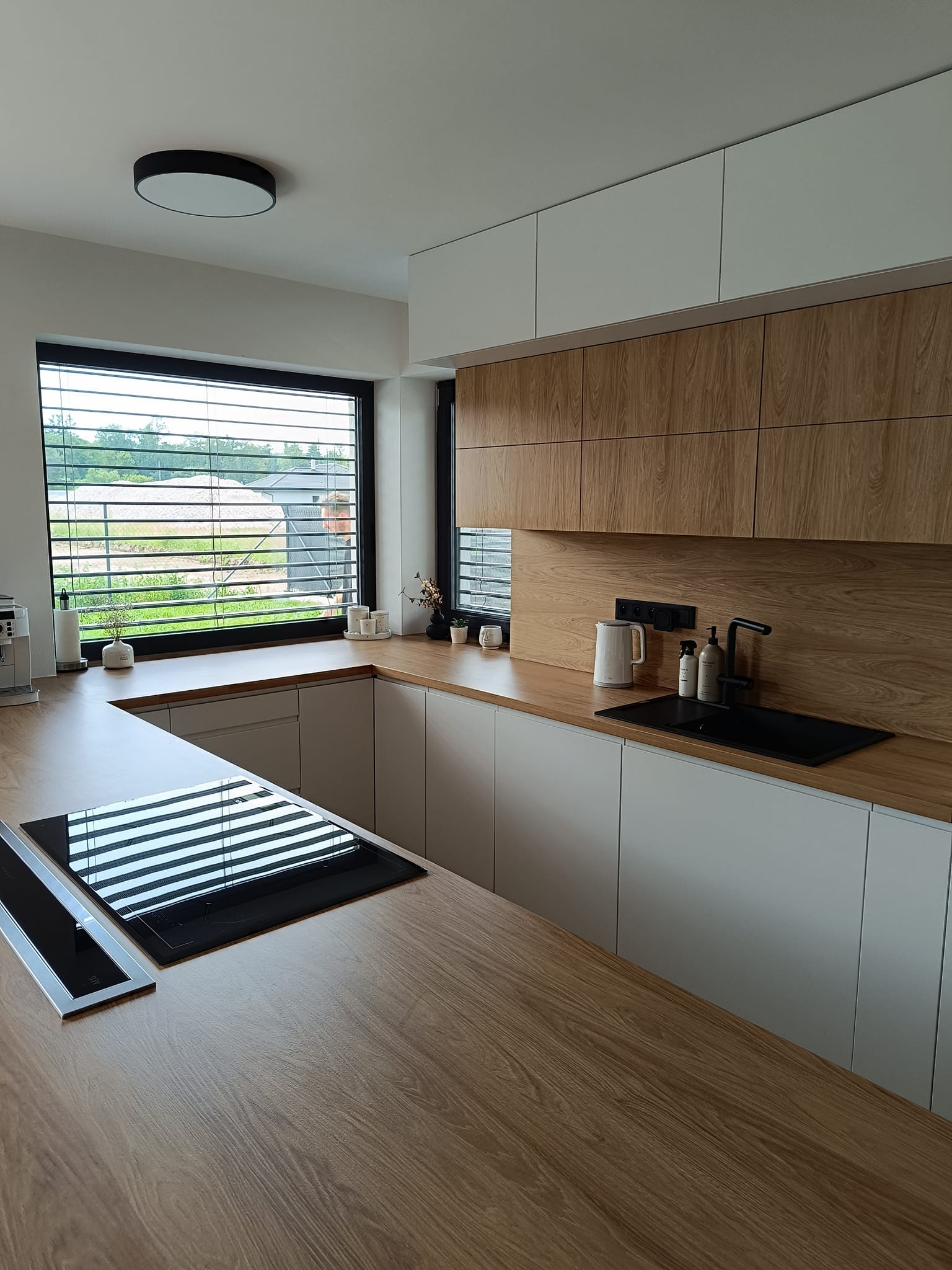 Nowoczesna kuchnia w odcieniach bieli i drewna z czarnymi akcentami: zlew, bateria, płyta indukcyjna. Minimalistyczny design, duże okno z żaluzjami.
