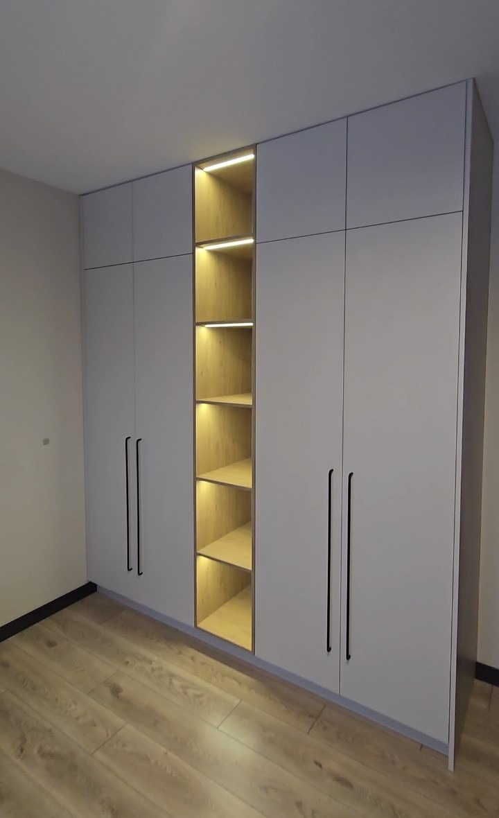 Jasnoszara szafa wnękowa z oświetloną wnęką na półki, czarne, długie uchwyty, drewniana podłoga. Minimalistyczny design.