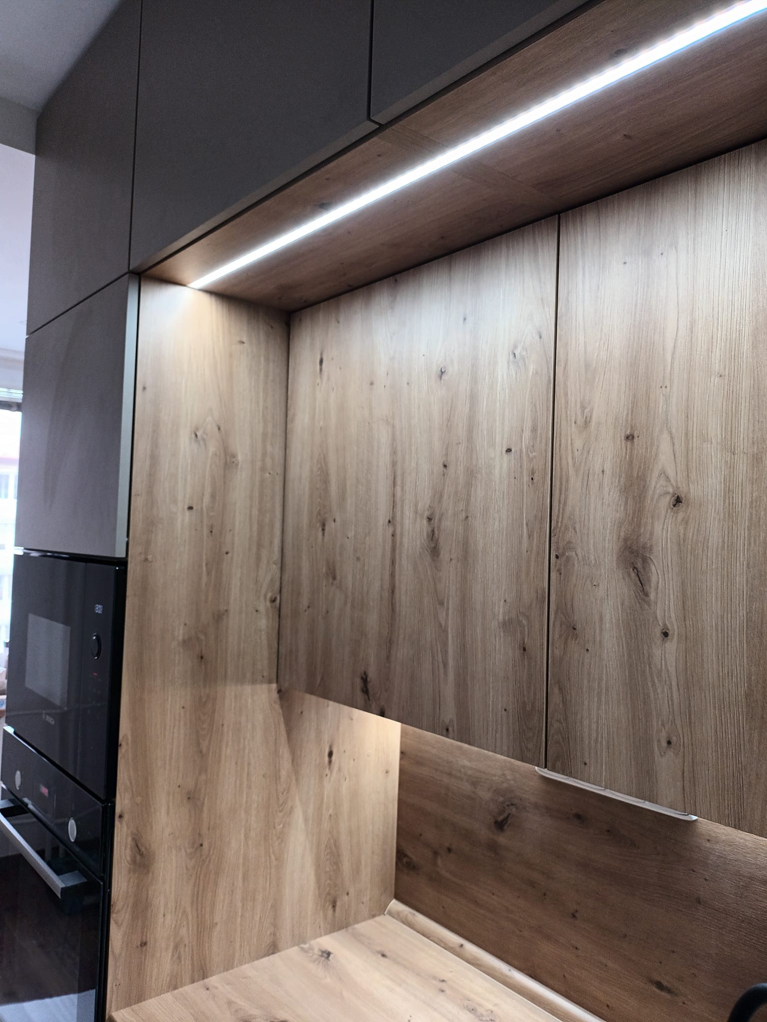 Drewniane fronty szafek kuchennych z oświetleniem LED, widoczny fragment zabudowy z piekarnikiem. Ujęcie z bliska, ukazujące strukturę drewna i detale wykończenia.