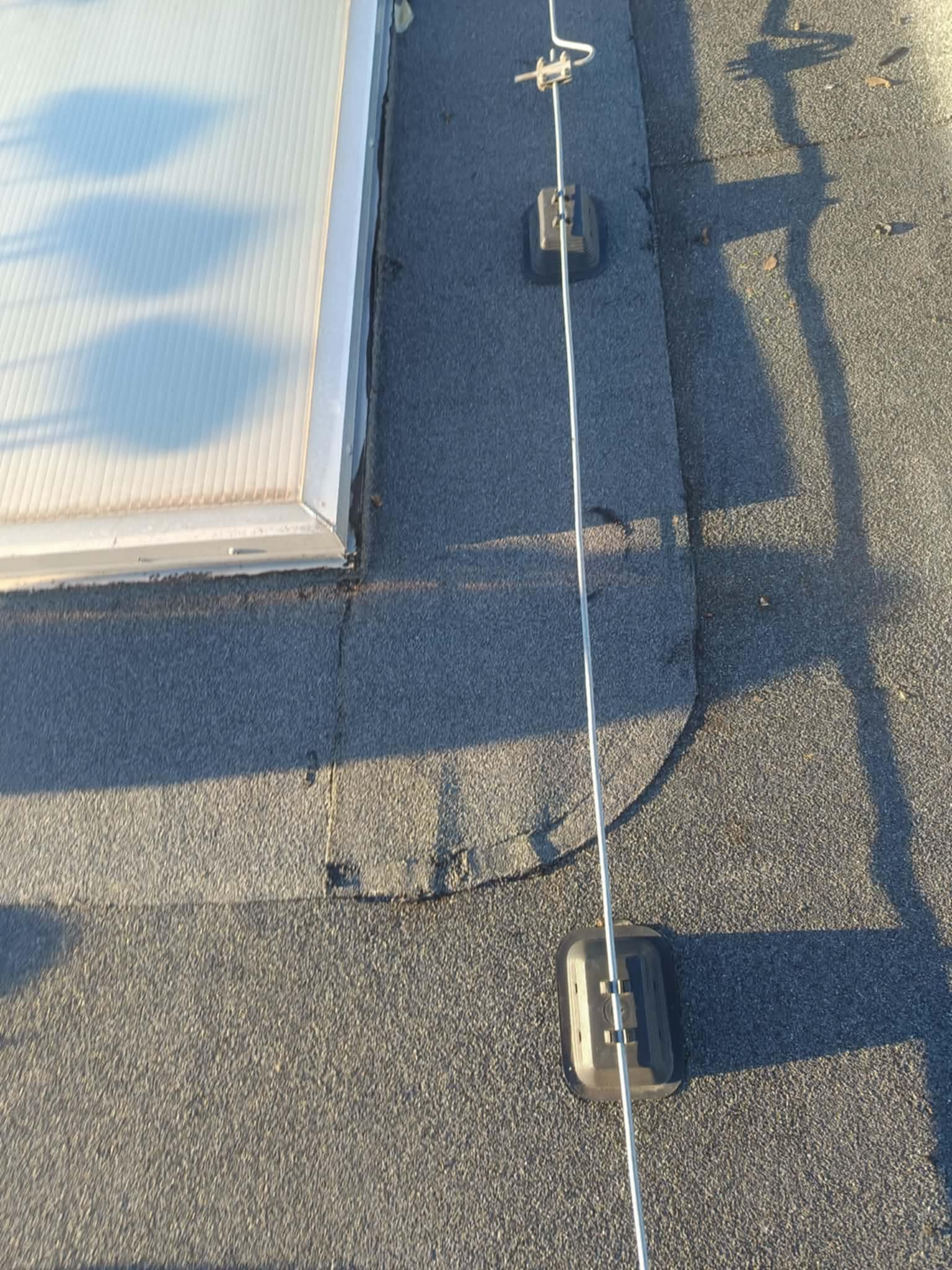 Fragment dachu pokrytego papą z zamontowanym świetlikiem i systemem odgromowym z uchwytami. Widoczne łączenia papy i cień.