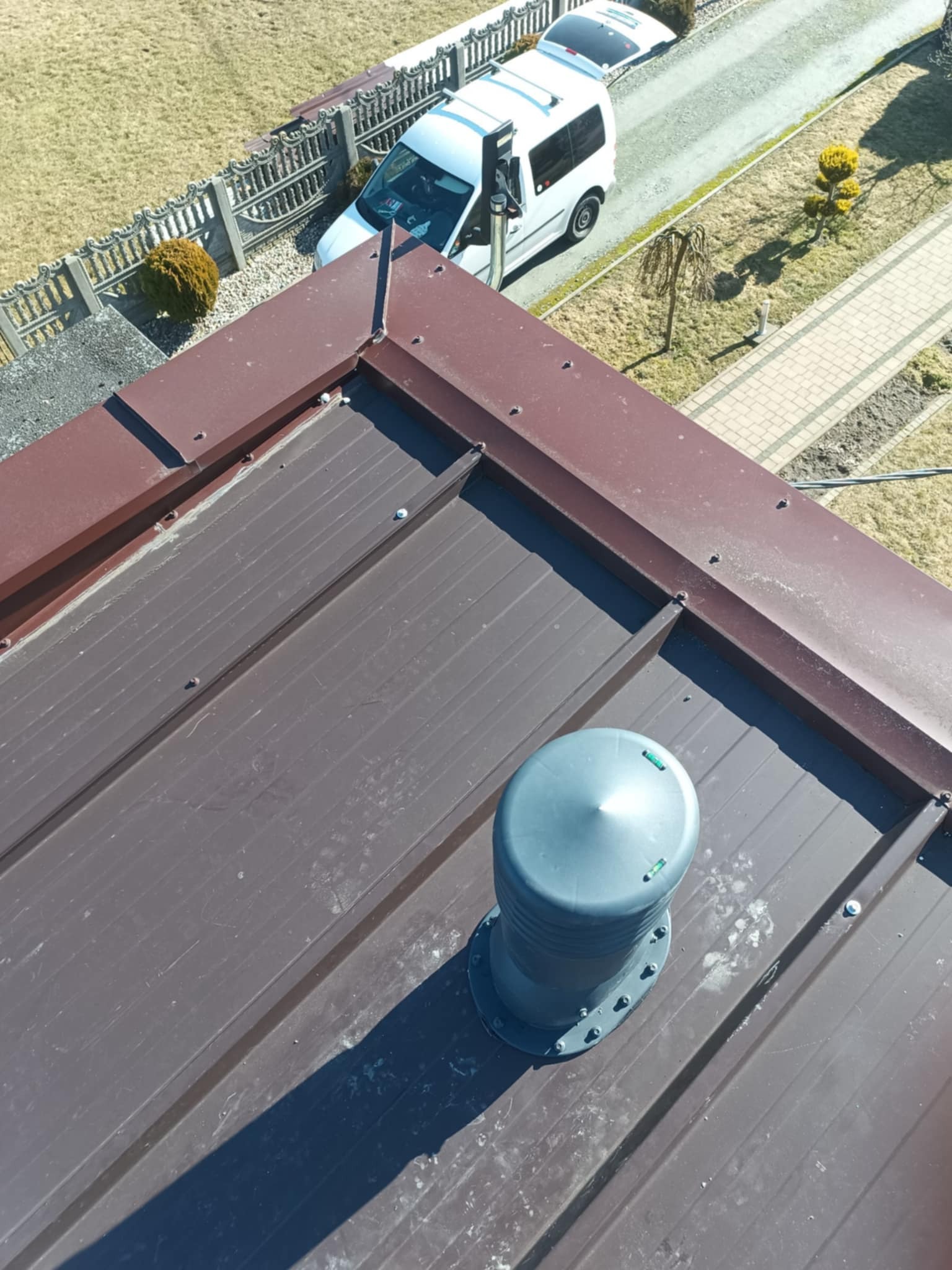 Brązowy dach z blachy trapezowej z zamontowanym odpowietrznikiem. Widok z góry na fragment dachu, zarys domu i zaparkowany samochód w tle.