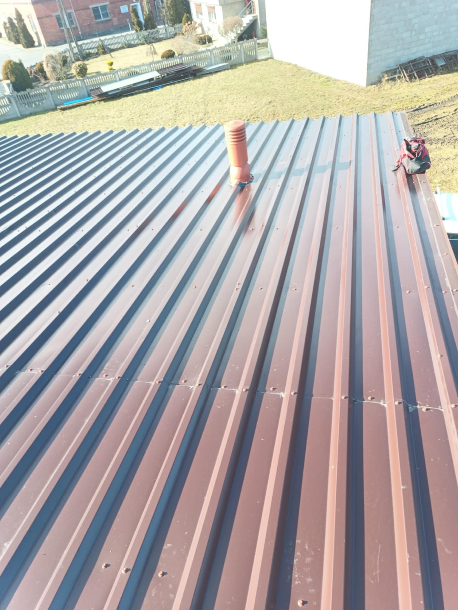 Nowy, brązowy dach z blachy trapezowej, widok z góry, z kominem wentylacyjnym i torbą narzędziową na krawędzi. Widoczne mocowania blachy. Trawa i budynki w tle.