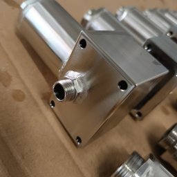 Tigstalmag - Precyzyjnie wykonane, polerowane elementy metalowe z gwintowanymi złączami, prawdopodobnie po obróbce CNC lub spawaniu, ułożone na brązowym tle.