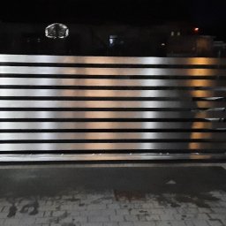 Tigstalmag - Nowoczesna brama wjazdowa z poziomych, metalowych profili, oświetlona nocą. Widoczny fragment ogrodzenia z szarej kostki i sylwetka mężczyzny w ciemnej kurtce.