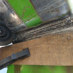 Tigstalmag - Szlifowanie spoiny na konstrukcji z zielonym elementem, zabezpieczonej drewnianą listwą i dociskanej ściskiem stolarskim. Widoczny fragment tarczy szlifierskiej i rękawica.